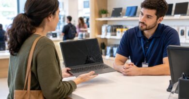 Mulher com bolsa marrom segura um notebook com tela rachada enquanto conversa com um atendente de camisa azul, em um balcão de loja com prateleiras ao fundo.