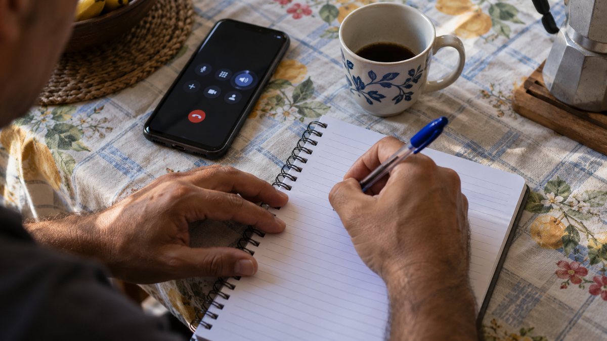 Mãos anotando o protocolo de atendimento em um caderno ao lado do celular em viva-voz